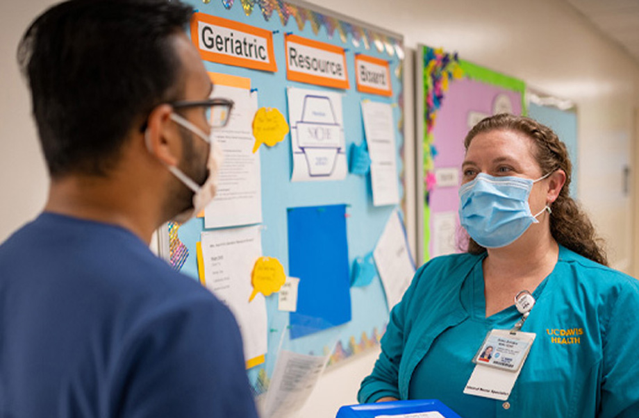 Nurse wearing scrubs and mask stands in front of bulletin board and talks to nurse colleague who looks at her and whose back is facing the camera